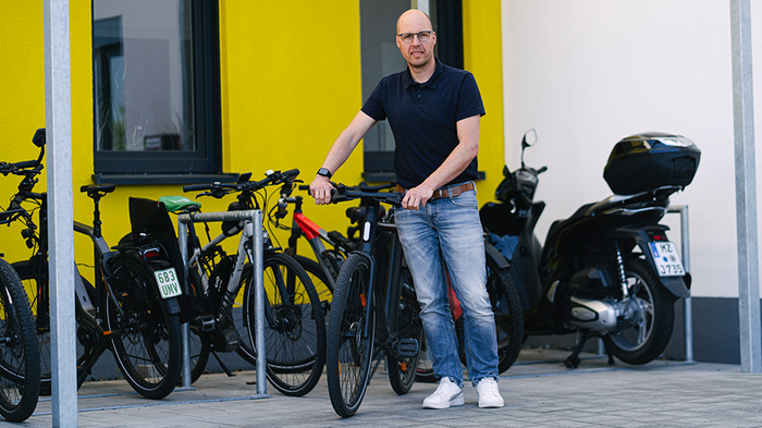 Mitarbeiter steht mit Fahrrad auf dem Unternehmensgelände vor einer gelben Wand 